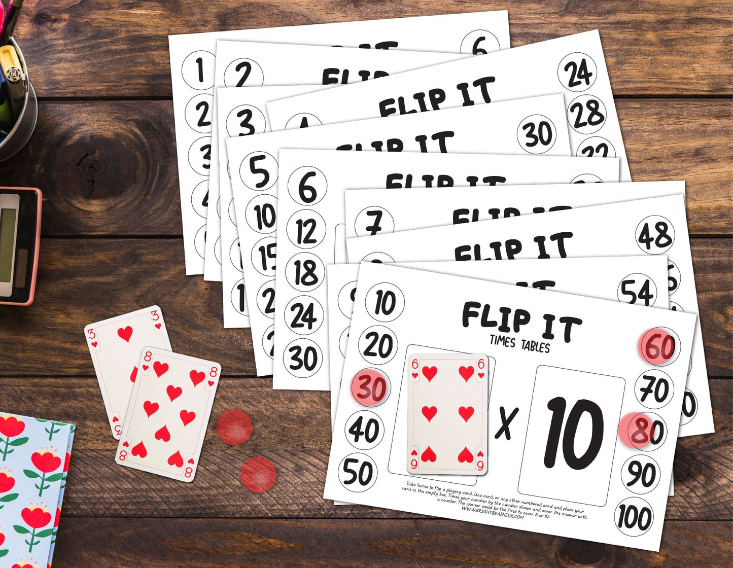 Flip it times tables. A set of educational math flashcards for times tables, including 'Flip It Times Tables' cards and playing cards, arranged on a dark wooden desk alongside a calculator, colored pencils, and a floral notebook.
